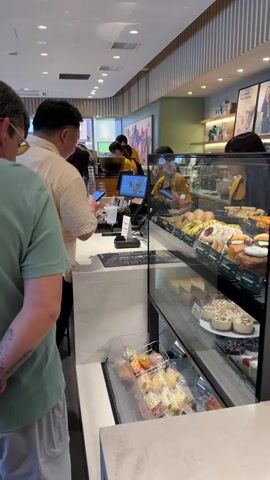 A man in a green t-shirt with tattoos on his forearms stands with his back to the camera, hands clasped behind him, waiting in a line. In front of him, a man in a beige short-sleeved shirt stands at a counter, appearing to interact with a female cashier in a yellow shirt who is operating a point-of-sale system with a computer screen and keyboard. To the right of the counter is a multi-tiered glass display case filled with various pastries, cakes, and individually wrapped food items like sandwiches or buns. The background features a light green wall with a large framed artwork and shelves stocked with merchandise. The ceiling has recessed lighting, illuminating the clean, modern interior of what appears to be a coffee shop or bakery.