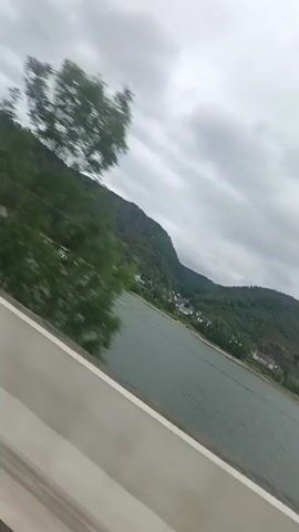 A landscape video taken from a moving vehicle shows a barge on a wide river. Mountains covered in green foliage rise steeply on the opposite bank, with a small village nestled at their base. The sky is overcast with grey clouds. The camera appears to be tilted, giving a dynamic perspective of the scene as the vehicle travels alongside the river.