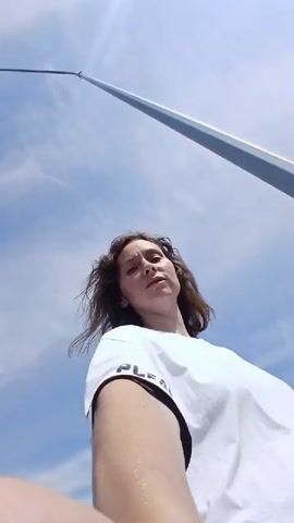 The video begins with a selfie-style shot of a woman with curly brown hair wearing a white t-shirt. She is outdoors, with a bright blue sky and scattered clouds overhead. To her left, a playground structure with orange and blue slides is visible, separated by a teal fence. As the video progresses, the woman turns slightly, revealing a young person with dark hair in the background, standing near the fence and looking away from the camera. Further back, cars are parked, and a line of trees forms the horizon. The woman continues to look at the camera and smile. The scene appears to be a public outdoor space, possibly a park or recreation area, on a sunny day.