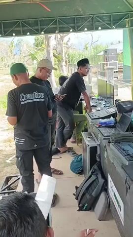 The video shows a group of people setting up for an outdoor event. There is a stage with a large screen and speaker system. Several individuals are working with audio equipment and computers, suggesting they are sound engineers or technicians. The environment appears to be a sandy area with some vegetation and trees in the background, under a cloudy sky. One person is wearing a t-shirt with the word "Creative" and a logo. The overall atmosphere seems to be one of preparation and organization for a public gathering.