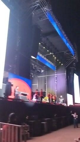This video captures a vibrant performance or concert scene at night or indoors. A large stage is set up with extensive scaffolding, numerous bright lights, and several prominent LED screens. On the stage, multiple individuals are engaged in what appears to be a choreographed performance or dance. One person is visible behind a console, possibly operating a sound mixer or DJ equipment. The lighting is dynamic, casting blue, purple, white, and red hues across the stage. The LED screens display various abstract patterns and colors. Crucially, one large screen prominently displays the text "FIESTAS FRÍAS" (Cold Parties) and "en Maipú" (in Maipú), confirming the event's name and its location in Maipú, Chile.