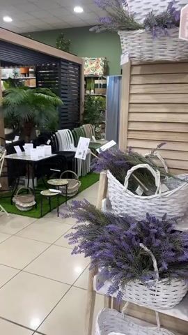 The video shows a retail store display with garden furniture under a pergola-like structure. Tables and chairs are arranged on artificial grass with decorative items like plants and baskets. The background features shelves with various home decor items. The scene is brightly lit with overhead lighting. The camera pans across the display showcasing the furniture and decorative elements.