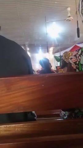 From a low angle, the camera is positioned behind a wooden pew in a church. A woman with dark hair and a colorful patterned headscarf and dress is seated in the pew in front of the camera. There is a book and papers in the pew's storage area. Other people are seated in the pews in front of her, but they are not clearly visible. The ceiling is white and has fluorescent lighting and a ceiling fan.