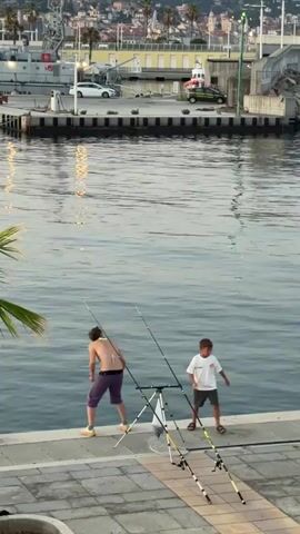 The video shows a harbor scene in Imperia, Italy, likely in the late afternoon or early evening due to the warm, soft lighting and the reflections on the water. Two people are visible on a paved walkway by the water's edge.

In the foreground, a person is fishing, holding a long fishing rod. They are wearing purple shorts and a bikini top, with their back to the camera. Two fishing rods are set up on a tripod stand. To the right, a young boy walks along the walkway, also facing away from the camera, wearing a white t-shirt and shorts.

Behind them, the harbor bustles with activity. Several boats are docked, including a large white vessel with a crane. A white car and a green vehicle are parked on the quay. Buildings with terracotta roofs are visible in the background, suggesting a Mediterranean coastal town. The water is calm, with gentle ripples and reflections of lights from the shore. The overall atmosphere appears peaceful and relaxed, typical of a seaside town at dusk. There is no visible text in the video.