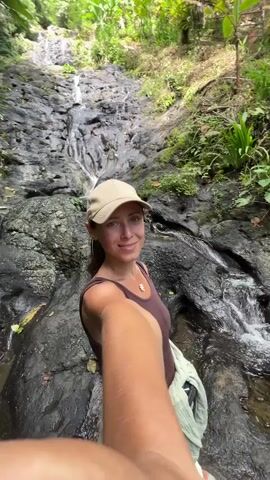A woman, wearing a brown sleeveless top and a tan baseball cap, is taking a selfie. She is smiling at the camera and winking. She has a green jacket tied around her waist, and a silver necklace with a pendant is visible. The background is a rocky slope with a waterfall cascading down it. Green foliage is visible on either side of the rocks. The woman's arm and hand are visible in the foreground, holding the camera. The setting appears to be a tropical or lush environment.