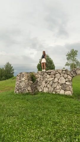 A young woman with long brown hair stands on a stone wall, looking down. She wears a dark blue crop top and a white skirt. The wall is made of large, rough stones and is covered in patches of green moss and grass. Behind her, a large green tree stands against a cloudy sky. The ground in front of the wall is covered in lush green grass with small white flowers. The camera zooms in on the woman as she walks along the wall.