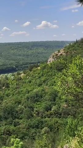 The video shows a panoramic view of a lush green valley with a river winding through it. The valley is covered in dense trees, with a mix of deciduous and coniferous species. The terrain is hilly, with steep slopes leading down to the river. The sky is partly cloudy, with patches of blue visible. There is no evidence of people or man-made structures in the immediate vicinity of the river, though a small village or cluster of buildings appears to be nestled along the riverbank in the distance. The overall impression is one of a serene and natural landscape.