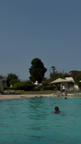 A man swims in a pool. In the background, there are lounge chairs, umbrellas, trees, and buildings.