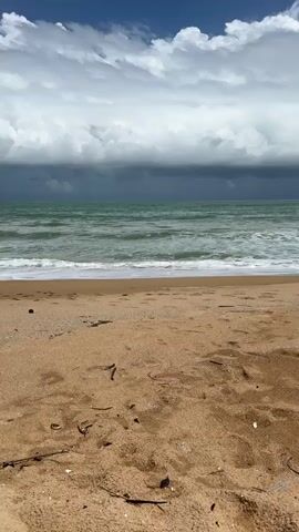 A static shot of a sandy beach with the ocean in the background. The ocean is a greenish-blue color with waves crashing on the shore. Above the ocean, the sky is a mix of bright blue with white clouds and darker, stormier clouds further in the distance. There are a few pieces of debris scattered on the sand in the foreground.