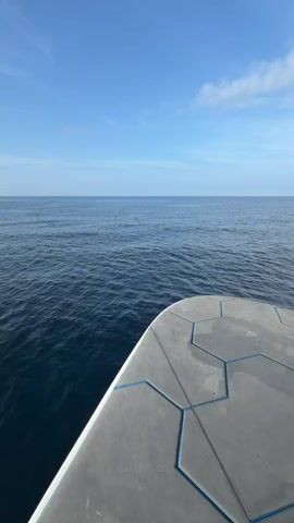 The video shows a still, eye-level shot from the front of a moving boat on a calm sea under a bright blue sky with a few wispy clouds. The boat's deck is visible in the lower portion of the frame, featuring a gray, textured surface patterned with blue hexagonal shapes. The vast expanse of the ocean stretches out to the horizon line, where the water meets the sky. The sea is a deep blue color with subtle wavelets rippling across its surface. The overall scene conveys a sense of tranquility and openness.