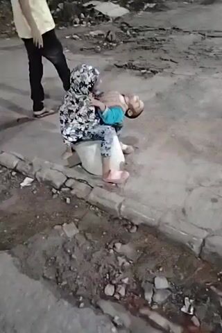 A child is held by another child. The child being held is lying on the other child's lap. The other child is sitting on a white object. There is debris on the ground and a curb. A person in a yellow shirt and dark pants is visible in the background.