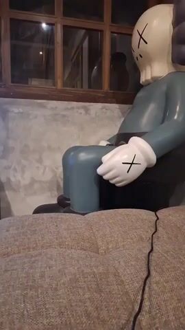The video begins with a close-up of a large, stylized sculpture, resembling a seated cartoon character with X's for eyes and a muted teal body. The sculpture is positioned on a textured gray couch or sofa in what appears to be an indoor setting. The background reveals a wooden-framed window with blinds, and the reflection in the glass shows an interior space with muted lighting and some objects, possibly furniture. The camera then pans to the right, shifting focus to a person's bare legs and feet, also resting on the couch. The person's right foot is visible in the foreground, and their legs extend further into the frame. The camera continues to pan, showing more of the window and the interior reflected within it. The scene is dimly lit, creating a relaxed or evening ambiance. There are no discernible actions or interactions occurring beyond the static display of the sculpture and the presence of the person's legs.