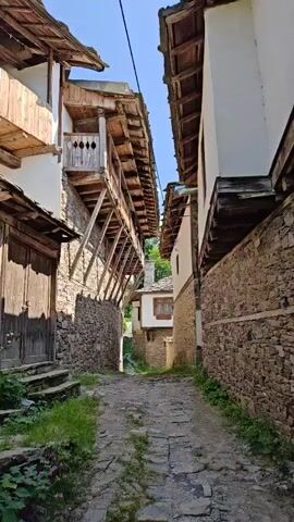 The video depicts two outdoor scenes, each of a cobblestone pathway surrounded by stone walls and traditional-style buildings. The first scene, at 00:00, shows a narrow pathway with steps leading up to a wooden door on the left. The buildings have wooden balconies and tiled roofs. The second scene, from 00:01 to 00:06, shows another narrow cobblestone path, this one with more vegetation growing alongside the stone walls. A utility pole is visible on the left side of the path. Both scenes are under a clear blue sky with some white clouds. There are no people present in either scene.