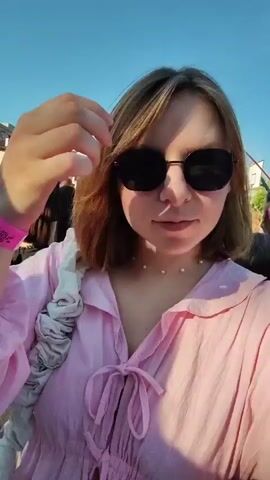 The video begins with a close-up of a young woman wearing a pink dress and sunglasses, her hand touching her hair. Behind her, a crowd of people are gathered outdoors, facing a stage where a performer is visible. The atmosphere suggests a concert or outdoor event. The woman smiles at the camera before the scene briefly shifts to a slightly wider shot showing more of the crowd and the stage setup. The sky is blue with some clouds. The woman then returns to the foreground, her hand still in her hair, as a pink balloon with a cartoon character on it floats into view from the right. The video ends with the woman continuing to touch her hair.