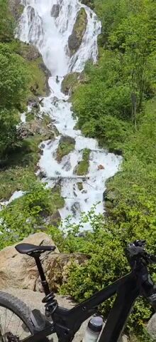 A person with dark hair runs down a very tall waterfall cascading over rocks, while two other people in bathing suits, one in yellow, stand on the side watching. A bicycle rests in the foreground. The waterfall is surrounded by green vegetation. The day is bright and sunny.