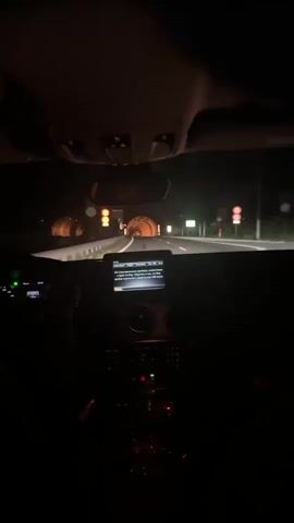 From the interior of a vehicle, the video shows the driver approaching a tunnel at night. The car's dashboard is visible, including the infotainment screen and the steering wheel. The interior is dark. Through the windshield, one can see the tunnel entrance, with lights illuminating its interior, and road signs on either side of the road. The driver is moving towards the tunnel.