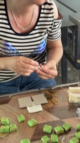 A woman is seated at a table, engaged in the preparation of small green sweets. She wears a black and white striped, off-the-shoulder top and blue jeans, accessorized with a silver necklace and a gold bracelet. Her expression is calm, with a gentle smile.

On the wooden patterned table in front of her are numerous small, cube-shaped green candies, which appear to have a textured, possibly nutty, surface. Adjacent to these are piles of small, clear plastic containers and stacks of square brown paper pieces. A piece of durian fruit with its distinctive yellow flesh is also visible on the table, to the right. A red plastic basket, partially seen, holds more of the green sweets.

The woman's hands are actively involved, appearing to be either unwrapping or preparing the sweets for packaging. Another hand briefly reaches into the frame to pick up some of the green sweets. The setting appears to be an indoor or covered workspace, likely a local confectionery or market stall where these traditional treats are made or prepared, consistent with the location of Thoi Son, Vietnam. No discernible text is visible in the frame.