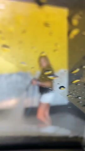 The video shows a young woman pressure washing a floor in what appears to be an outdoor covered area. The background features a vibrant yellow wall, a concrete barrier, and a blue hose. The woman is wearing a black top with lace detailing and white shorts. She is holding a pressure washer and directing a spray of water and foam at the ground. The lighting suggests it is daytime. The setting could be a car wash or an area designed for cleaning, given the presence of the pressure washer and the wet ground. The woman's expression is neutral as she focuses on the task. There are no visible signs or text in the video. The location is identified as Mišići, Montenegro.