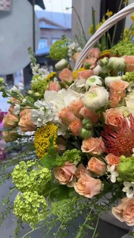 The video shows a flower arrangement in a white basket with a handle. The arrangement includes a variety of flowers in shades of peach, white, yellow, and green. Roses, ranunculus, chrysanthemums, hydrangeas and other greenery are visible. The basket is lined with white tissue paper and has long white ribbons. The background shows a window with a view of buildings and trees. There are also rolls of colorful ribbon visible near the right edge of the frame.