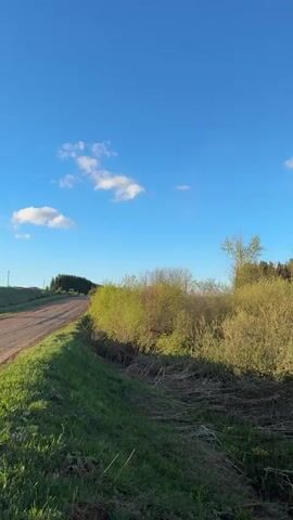 The video shows a dirt road in a rural setting, with green grass and vegetation along the roadside. The sky is blue with scattered clouds. The camera pans across the landscape to reveal a distant horizon with rolling hills. The sun is partially obscured by clouds, creating a bright glare. There are power lines and utility poles visible against the sky.