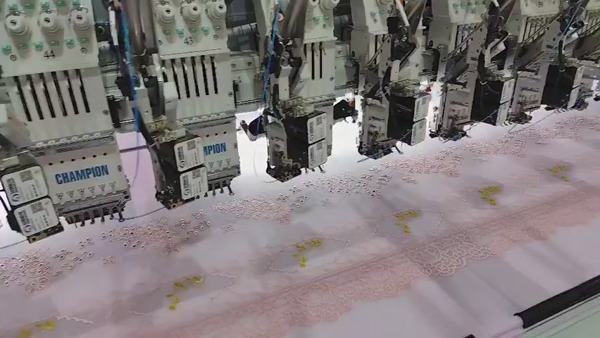 This video shows an industrial embroidery machine with multiple sewing heads working in unison on a large piece of fabric. The machine is very long, with at least ten sewing heads visible in a row, each attached to a vertical support structure. The sewing heads are silver and white, with various mechanical components and numerous spools of thread at the top. Each head has a black box-like component near its base. The fabric being embroidered is light pink and appears to be delicate, possibly lace or a similar material. The embroidery itself consists of small, intricate patterns in pink and possibly yellow thread. The lighting is bright, indicating an indoor environment, likely a factory or trade show. In the background, a large sign with green lettering that says "AMPION" is visible. Near the far right of the frame, a man wearing glasses, a white polo shirt, and a lanyard with a badge is standing and observing the machine. The camera angle is high and slightly angled, looking down at the machine and the fabric. The overall impression is one of automated textile production.