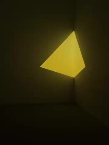 Okay, here's a description of the video:

The video shows a corner of a room, possibly an art installation, illuminated with a bright yellow, triangular light. The corner is formed by two walls that meet at a 90-degree angle and the floor. The light source itself is not visible, but the triangular shape of the light suggests it may be coming from a triangular opening or a light fixture shaped that way, positioned behind the walls. The surrounding walls are dark, which intensifies the effect of the bright yellow light.