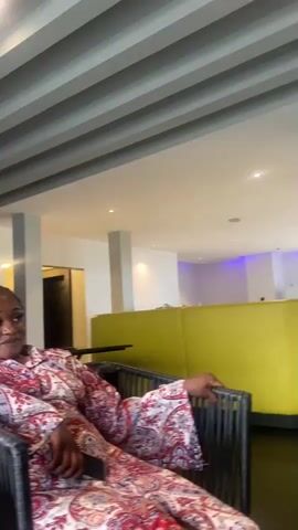 The video shows a woman sitting in a chair in what appears to be a lounge or restaurant. She is wearing a patterned dress. Behind her, there is a bar area with shelves of bottles and glasses. Another person is visible behind the bar, facing away from the camera. The ceiling has a slatted design. The camera is moving slightly throughout the video.