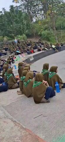 A large group of young people, likely students or scouts, are seated on tiered concrete steps outdoors. They are mostly wearing brown uniforms with green scarves. Many are wearing hijabs. Some are holding colorful pinwheels. The environment appears to be a park or open-air amphitheater, with trees and a building visible in the background. The people are facing forward, suggesting they are watching an event or listening to a speaker. The overall atmosphere seems calm and attentive.