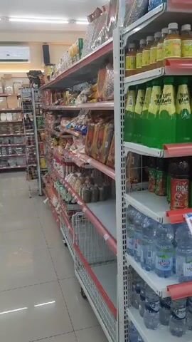 The video shows an interior view of a well-stocked convenience store or supermarket aisle.  Shelves are filled with various packaged goods, including snacks, drinks in bottles, and other food items.  The lighting appears to be fluorescent and overhead.  The camera is moving, panning from left to right, offering a perspective of walking down the aisle.  There are no people visible in the frame.  The shelves are made of metal with red trim.  The floor is tiled.