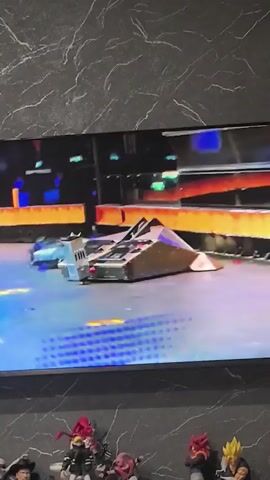 A television screen displays a robot combat competition. Two robots, one larger and silver with a sloped front, and a smaller, blue robot with a different shape, are in the arena. The arena floor is blue and purple with yellow circular markers. The robots are moving and engaging in combat. Flames are visible along the arena walls. In the foreground, below the television, are several collectible figurines of anime characters.