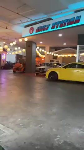 The video shows several parked cars under an overhang labeled "Juicy Station." The cars include an orange jeep, a silver sedan, a yellow coupe, a white hatchback, and a gray coupe.  The area is lit with string lights and ceiling lights, and the floor is gray concrete. The overhang appears to be part of a building or structure. The camera slowly pans from left to right, showcasing the cars parked.