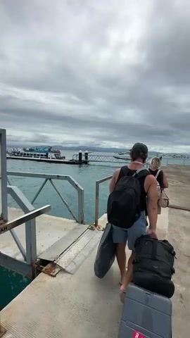 A man pulls a suitcase and carries two bags on a pier under an overcast sky with a view of the ocean, boats, and a distant shoreline. Another woman walks ahead of him. The pier has metal railings on both sides and appears to be made of concrete with some metal components. The water is a light blue-green color.
