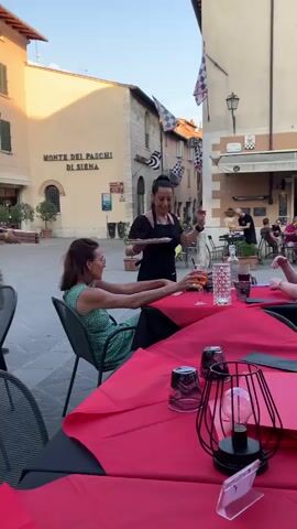 The video shows a scene unfolding in an outdoor dining area in San Quirico d'Orcia, Italy. Several tables are set with bright red tablecloths, suggesting a restaurant or cafe.

In the foreground, two women are seated at a table, engaged in conversation. One woman, wearing a green dress, is looking towards the server. The other woman, in a blue dress, is seated opposite her. A waiter, dressed in an apron over a black outfit, approaches the table, holding a pizza. Another waiter, in a tan shirt and black apron, is visible in the background near a bar area.

The background reveals a charming Italian piazza. A building with the sign "MONTE DEI PASCHI DI SIENA" is prominently visible. The architecture suggests a historic Italian town. Flags with checkered patterns are displayed, adding a festive touch. The warm, golden light indicates it is late afternoon or early evening, with pleasant weather conducive to outdoor dining.

The overall atmosphere appears relaxed and convivial, with people enjoying a meal and each other's company in a picturesque setting. The visible text on the building is "MONTE DEI PASCHI DI SIENA".