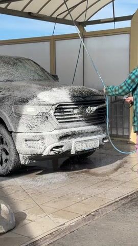 A male individual is captured in an outdoor self-service car wash bay in Bezborodovo, Russia, during what appears to be a bright, sunny day. He is seen washing the front of a black RAM pickup truck, which is heavily covered in white soap foam.

The man, positioned on the right side of the frame, is wearing a green and blue plaid jacket over a dark hoodie, and purple pants. He is actively using a high-pressure washer, holding the spray wand and directing a stream of water onto the foamed truck, specifically targeting the grille area to rinse off the soap.

The truck's prominent "RAM" emblem is clearly visible on its grille, despite being partially obscured by foam. The front license plate is also discernible, displaying the characters "C570CC 99", with "99" being a Russian region code, likely for Moscow.

The car wash bay features a light-colored paved floor, a metal-framed canopy overhead providing partial shade, and translucent wall panels on the sides. A small concrete barrier is visible in the lower-left foreground. The overall scene depicts a routine vehicle maintenance activity.