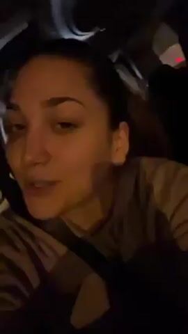 A young woman is sitting in a car at night. She is wearing a beige hoodie, and her hair is pulled back in a ponytail. She is singing along to a song playing in the car, and she looks into the camera, occasionally turning her head slightly to the side. The interior of the car is dark, but lights can be seen through the window. She seems relaxed and casual.