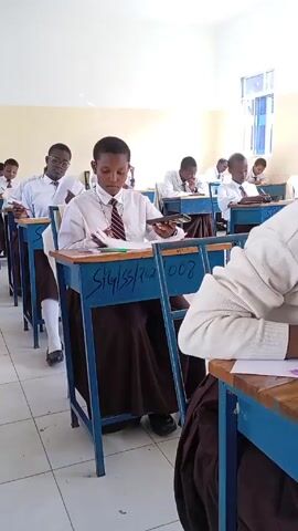 Okay, here's a detailed, matter-of-fact description of the video:

The video shows a classroom full of students who appear to be taking an exam. The students are seated individually at blue desks arranged in neat rows. They all wear white shirts, brown skirts or pants, and ties. The camera angle shifts during the video, offering different views of the classroom.

Some students are actively writing on paper, while at least one is holding a phone, looking at it intermittently between writing. Other objects on the desks include pens, pencils, erasers, and a mathematical set. The classroom has white walls, and natural light is coming through a window visible in the background.  The overall impression is one of a formal, academic setting during an examination period.