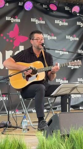A man is sitting on a folding chair on a stage and playing an acoustic guitar. He is wearing a black collared shirt with stripes and black pants. He has on glasses and is singing into a microphone. Behind him is a black banner with pink butterflies and the words "BAKgarden" repeated. There are lights shining on him from above. In front of him, there are speakers and grass. A water bottle is on the floor near his feet.