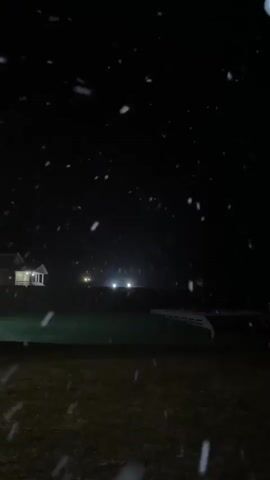 The video depicts a dark outdoor scene at night with snow falling. A lawn is visible in the foreground. In the distance, there is a house with a light on and what appears to be street lights. The falling snow is highlighted against the dark background. There is no interaction or people visible. The scene is primarily focused on the snow.