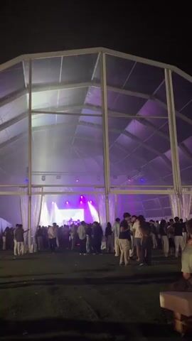 The video shows a large, clear-span tent structure with a geodesic dome-like roof. Inside the tent, a stage is set up with a DJ booth and lighting equipment. The stage is illuminated with bright purple and white lights, creating a vibrant atmosphere. A crowd of people, dressed in casual attire, are gathered in front of the stage, some dancing and socializing. Outside the tent, the ground is covered in grass, and the night sky is visible above. The overall scene depicts an outdoor music event or party.
