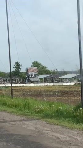 The video shows a rural scene from the perspective of a moving vehicle. Initially, damaged buildings, including what appear to be agricultural structures with partial roofs, are visible beside a dirt road. There are utility poles and lines present. The landscape includes a field with planted rows. As the video progresses, the vehicle travels along a paved road with cracks and patches. A series of tall poles connected by netting stretches over the road, creating a tunnel-like effect. The surrounding environment consists of grassy areas, trees, and glimpses of buildings in the distance. There are signs alongside the road, and another vehicle can be seen ahead.