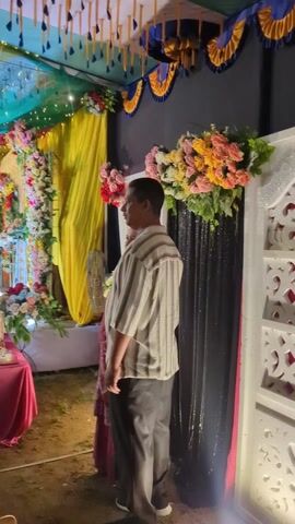 The video showcases a brightly decorated event space, possibly a wedding or celebration. The scene features colorful draped fabrics, floral arrangements, and stage lighting. People are visible, including photographers adjusting equipment and individuals posing for photos. A small table holds what appears to be a cake and other food items. The overall atmosphere is festive, with attention focused on capturing memories.