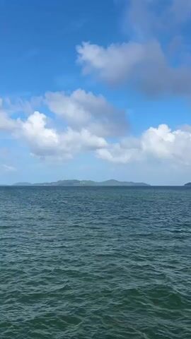 A vast expanse of deep blue ocean stretches across the lower half of the frame, its surface rippled by gentle waves. Above, a bright blue sky is dotted with fluffy white clouds. In the distance, a chain of islands with green, tree-covered hills forms a distant horizon. On the left side of the frame, a wooden pier juts out into the water. A small white boat is docked at the pier, and a construction crane stands nearby.