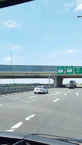 The video shows a highway scene from inside a vehicle. A white car is driving in the middle lane. To the right, there is a truck in the right lane. Overhead, there are green road signs indicating directions for A2 Bucuresti, A2 Cartier Titan, and A0 Pitesti Oltenita. The road has white lane markings. The sky is blue with some clouds, and the overall environment appears to be daytime on a sunny day. There is an overpass above the highway.