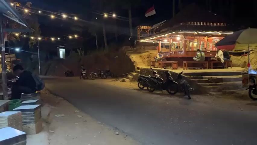 The video captures an outdoor scene at night. A road runs through the foreground, with a few motorcycles parked on the side. On the right side of the road, there is a food cart under a colorful umbrella. Across the road, an elevated wooden structure, possibly a restaurant or cafe, is illuminated by string lights. Several people are seated at tables within or around this structure. A set of stairs leads up to the structure. In the background, trees are visible, and an Indonesian flag is flying from a pole above the structure. The overall ambiance suggests a roadside eatery or gathering place at night.