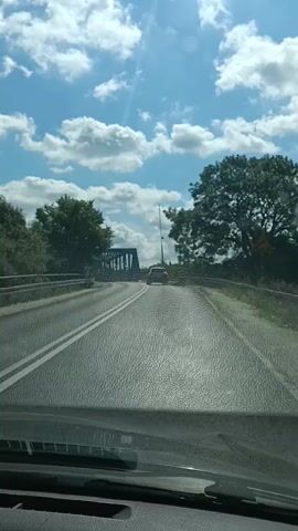 This video captures a perspective from inside a vehicle driving across a bridge. The road surface is visible, marked with white lines. The bridge appears to be constructed with a metal framework, and a smaller vehicle is seen ahead. On the left side of the road, there's a guardrail and lush green trees. On the right, a larger tree stands prominently, and beyond that, a red barrier is visible. The sky is a vibrant blue, dotted with fluffy white clouds, suggesting it's a sunny day. In the distance, a tower-like structure is faintly visible. The overall scene depicts a scenic drive on a clear day, likely in a rural or semi-rural setting. The time of day appears to be daytime, given the bright sunlight. No text is visible in the video.