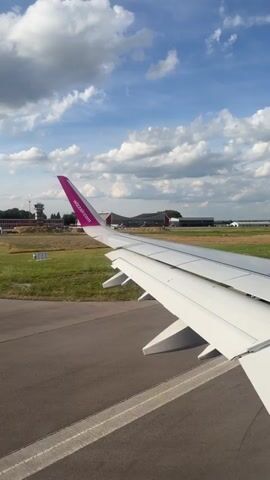 This media file shows a view from an airplane window, looking out over the wing. The airplane is on a runway, with buildings and a control tower visible in the distance. The sky is blue with white, fluffy clouds, suggesting fair weather. The overall scene is one of an airport in daytime. The prominent object is the airplane's white wing, with its distinctive purple tailfin featuring the text "Wizzair.com". The runway is asphalt, with white markings. Green grass surrounds the runway, indicating a rural or suburban airport setting. There are no people visible, and the focus is on the aircraft and its surroundings. The activity appears to be the moments before takeoff or after landing.