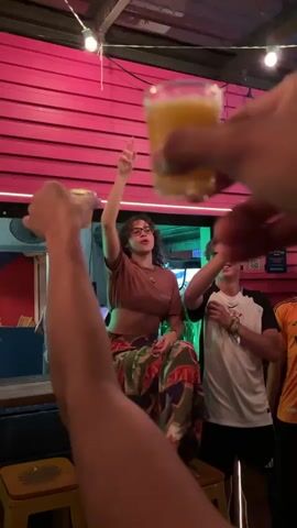 The video depicts a lively scene inside a bar or casual restaurant, characterized by vibrant pink slatted walls and neon green and yellow lighting. Several people are engaged in a celebratory activity, most likely reacting to a sports event.

In the foreground, a person's arm and hand holding a drink are raised in a toast or cheer. Further back, a woman with curly hair and glasses, wearing a brown crop top and patterned pants, is seated on a stool, expressing excitement. She is initially pointing upwards, then laughing and covering her mouth. Next to her, a man in a white and black sports jersey, possibly a Corinthians shirt, is also holding up a drink and cheering. The background shows parts of a bar counter and another man.

Throughout the video, a group of people can be heard loudly cheering and chanting "Dois! Um! Gol!" (Two! One! Goal!), strongly suggesting they are watching a soccer match and celebrating a goal. The overall atmosphere is enthusiastic and joyous. The time of day appears to be evening or night due to the indoor lighting. A partially visible sign on the wall displays a phone number, "305-303-769," which is a US area code.