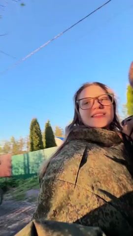 The video shows a woman with glasses wearing a green camouflage jacket sitting behind a man on some kind of vehicle. The man is wearing a black hoodie. The camera is pointing up at the couple, capturing the blue sky and a few houses and trees in the background as they move along a road. The sun is shining.