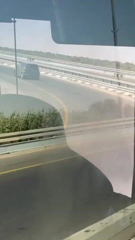 The video captures a view from inside a vehicle, likely a bus, looking out at a highway in the Bnei Shimon Regional Council, Israel. The day appears bright and sunny, suggesting fair weather. The scene is dominated by a multi-lane highway curving ahead, with a dark-colored SUV traveling on the upper lane. In the foreground, a yellow line marks the lane of the vehicle from which the video is recorded, with a guardrail separating it from the upper highway. A sign with black and white chevron arrows indicating a curve is visible. Beyond the highway, sparse vegetation and a few distant power lines are apparent. Reflections on the window obscure parts of the view, adding a layered effect to the imagery. A white bus can be seen on the upper highway in some frames, and a dark SUV is consistently visible. The overall impression is a snapshot of travel on a sunny day along an Israeli highway.