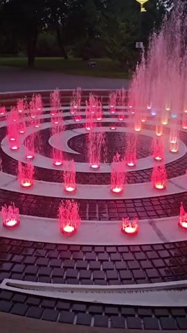 This video captures a dynamic fountain display at dusk in Keila, Estonia. The fountain is arranged in concentric circles, with numerous jets of water erupting from illuminated cylindrical bases. The lights beneath the water create a vibrant spectacle, predominantly in shades of pink, with hints of orange and yellow visible in the further rings. The water itself is pinkish, reflecting the colored lights.

The setting appears to be a park or public square, with mature trees silhouetted against the darkening sky in the background. A single, modern lamppost with a yellow glow is visible in the distance. The ground around the fountain is paved with dark square tiles, bordered by circular bands of lighter material. The overall atmosphere is serene and visually captivating, suggesting a pleasant evening in a well-maintained urban or park setting. No people are visible in the frame, and there is no visible text. The time of day is clearly evening, as indicated by the low light conditions and the artificial illumination of the fountain.